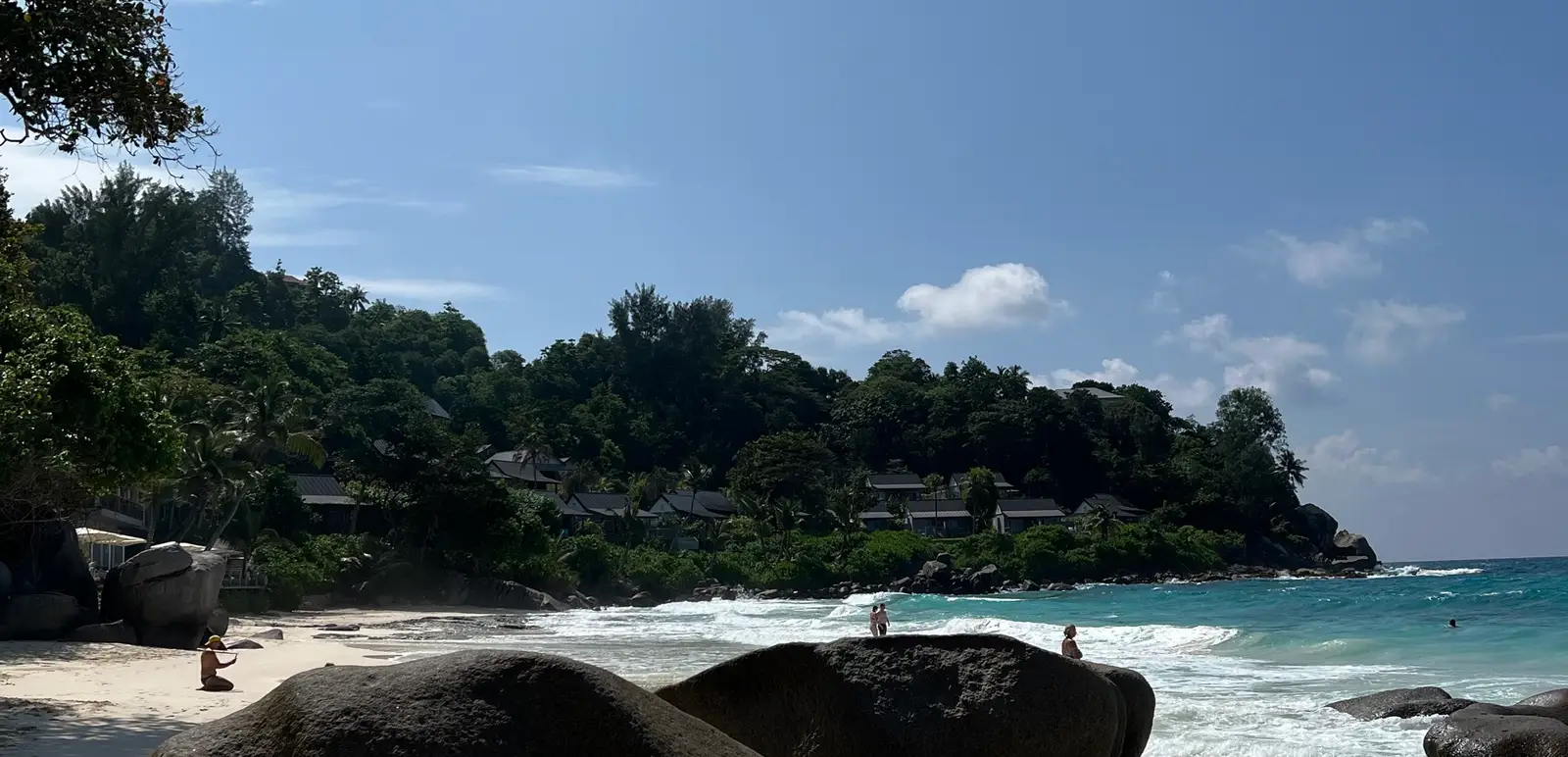 Seychelles beach lined with large rounded pink and grey granite boulders in the foreground, bathers' silhouettes in the turquoise lagoon and tropical vegetation behind.
